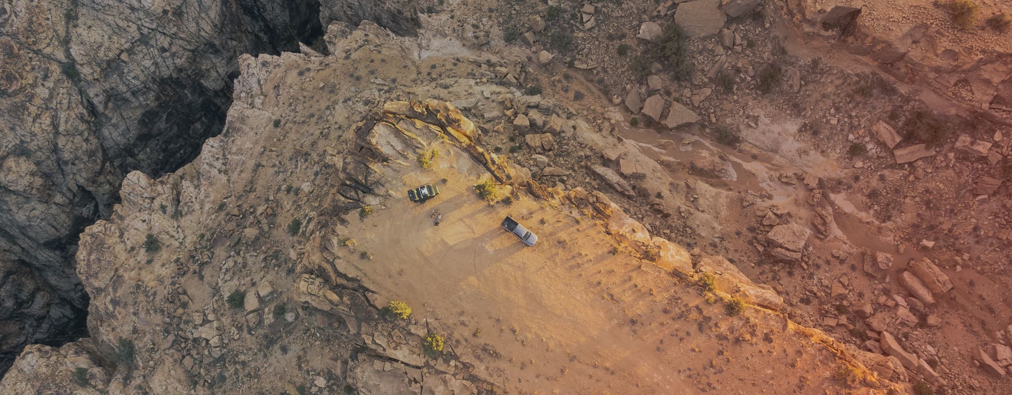 Aerial view of an isolated campsite on the edge of a dramatic canyon, with two off-road trailers and vehicles parked near a fire pit.