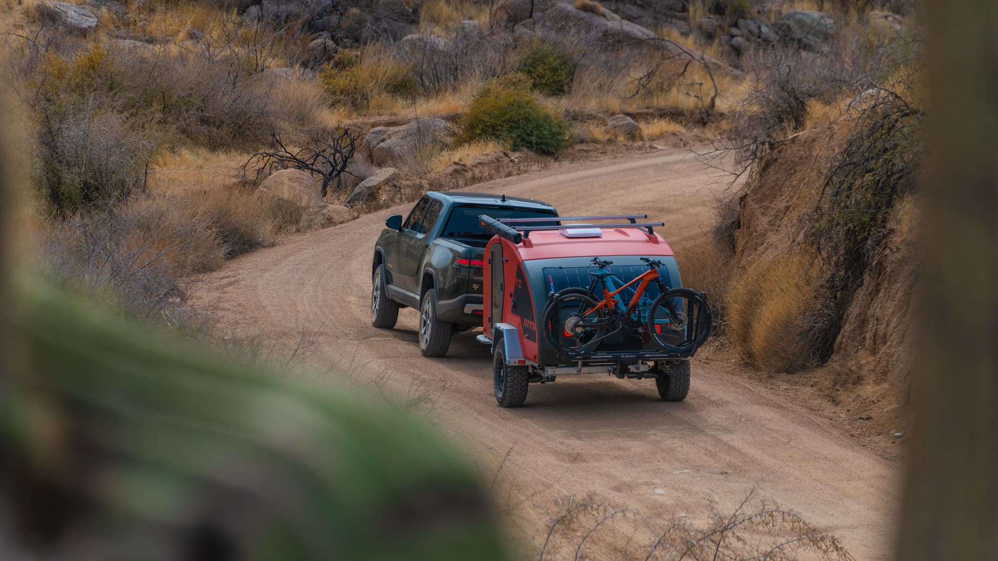 Rear trailing shot of a Rivian towing the Escapod TOPO2 MTB trailer with two mountain bikes mounted, curving through a dusty desert canyon trail.
