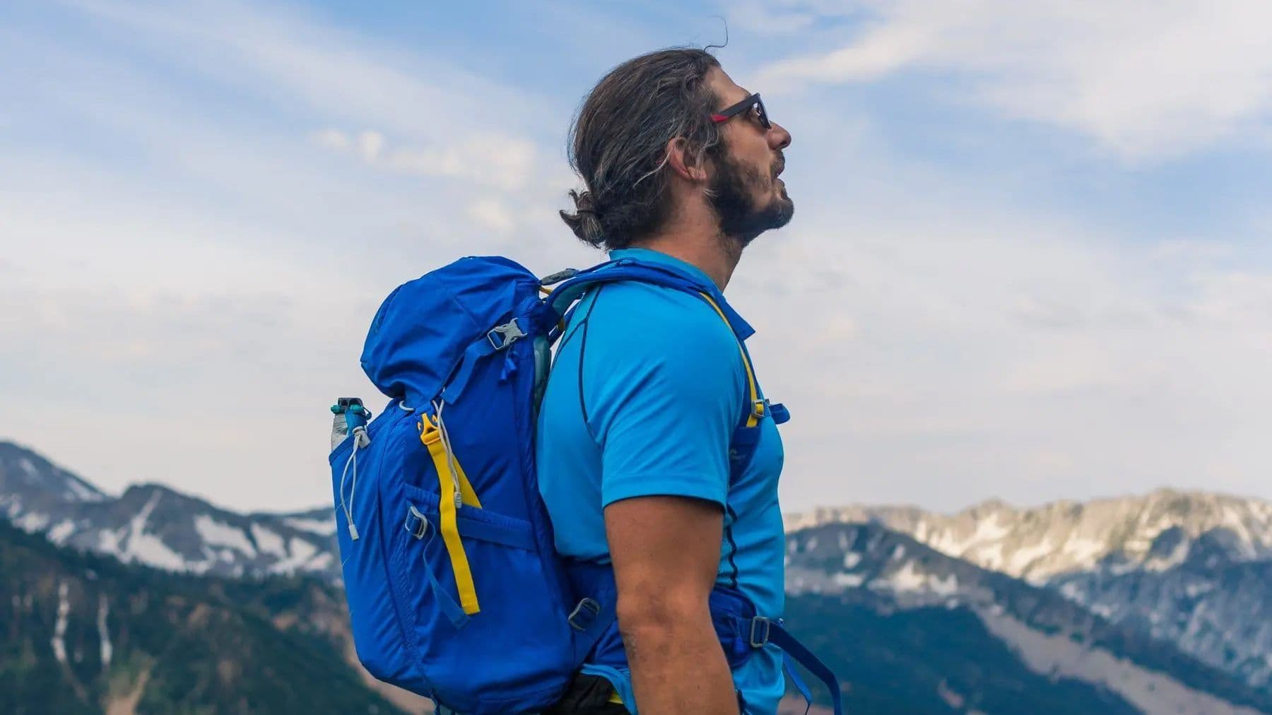 A bearded man in a bright blue hiking shirt and sunglasses stands with a blue backpack, gazing up at the sky with a mountainous landscape behind him.