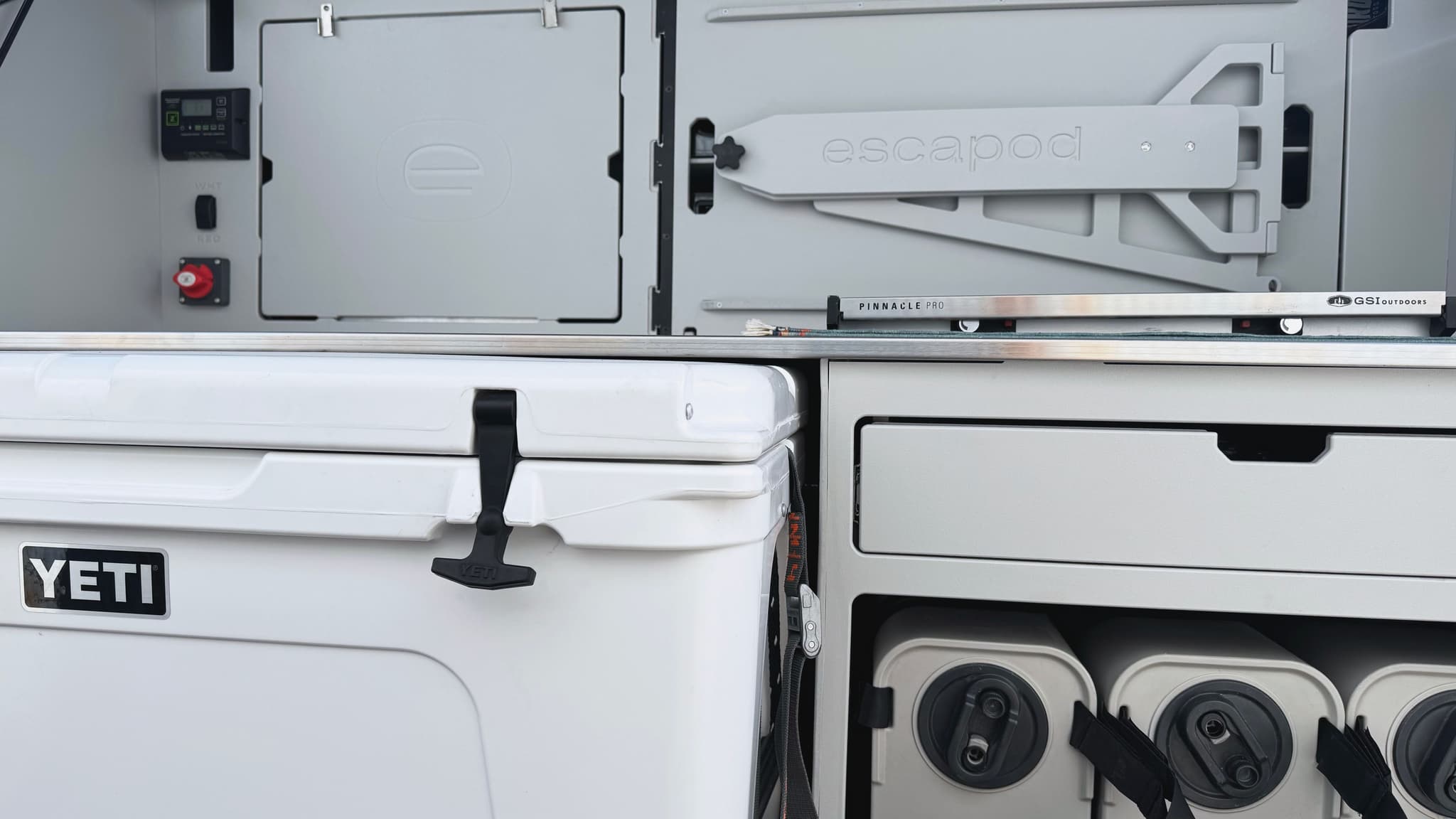 A low, wide angle view of the Escapod TOPO2 Nomad galley, showing a white YETI cooler, three Dometic water jugs, built in cabinetry and drawers made out of HDPE, and a wide stainless steel countertop.