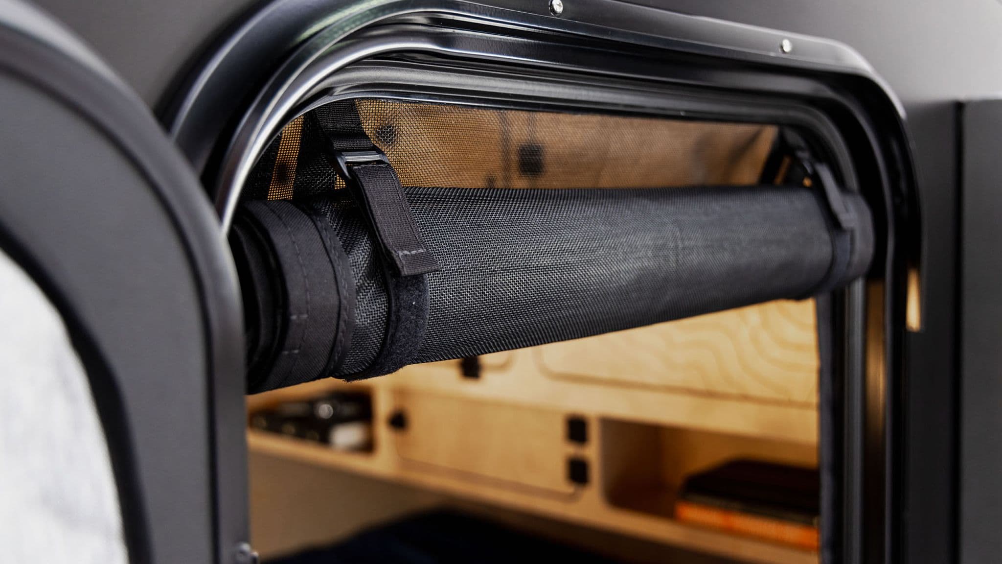 A close-up of the retractable bug screen rolled up inside the doorway of an Escapod TOPO trailer, revealing a cozy wooden interior.